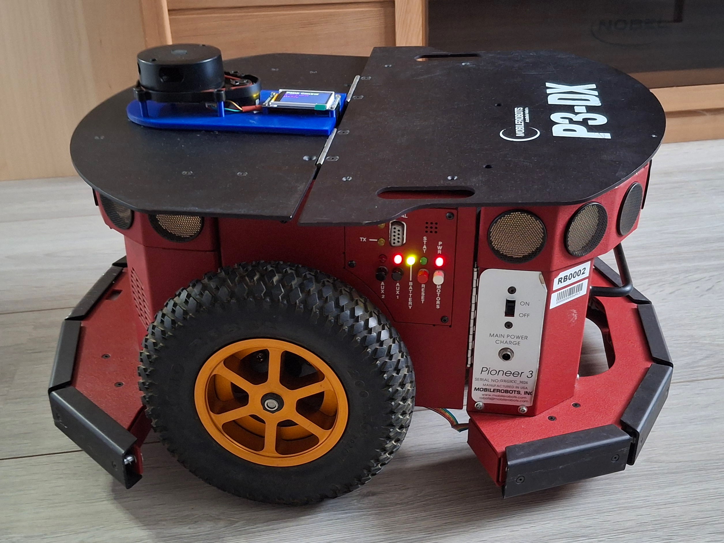 Pioneer 3DX mobile robot with red chassis and black top deck displaying P3DX branding, equipped with mounted LIDAR sensor on top platform and 1.77 inch status display on front panel, featuring yellow-rimmed wheels with rugged tires, positioned on light wooden floor against wooden furniture background